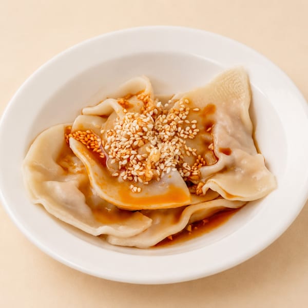 A white bowl filled with dumplings drizzled in chili oil and topped with sesame seeds.