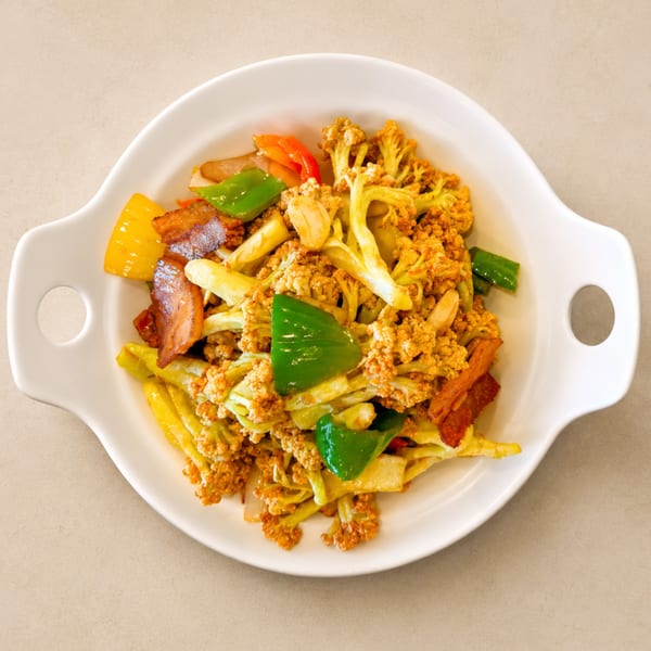 Stir-fried cauliflower with bacon, green and yellow bell peppers, and garlic in a white dish.